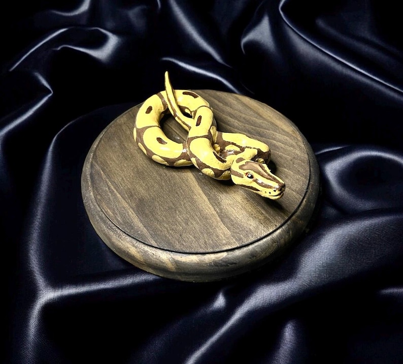 May include: A detailed, handcrafted figurine of a yellow and brown snake coiled on a round, wooden base. The snake's scales are meticulously painted, and the base has a dark, weathered finish. The background is a dark, silky fabric.