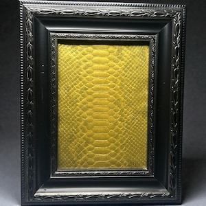 May include: A black rectangular picture frame with decorative detailing. Inside the frame is a textured, golden-yellow surface with a snakeskin-like pattern. The frame has a dark, matte finish.