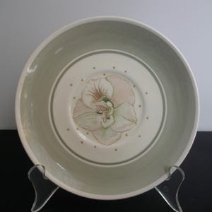 May include: A decorative ceramic plate with a floral design. The plate features a central image of a flower in shades of pink, green, and white, surrounded by a light green border. Small gold dots encircle the flower.