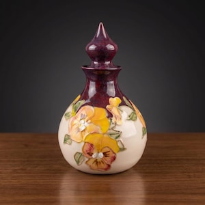 May include: A ceramic decanter with a bulbous base and a pointed stopper. The upper portion and stopper feature a deep, glossy burgundy glaze, while the lower body is cream-coloured and decorated with raised, hand-painted yellow pansy flowers.