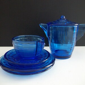 May include: A cobalt blue glass tea set with a teapot, cup, and saucer. The set is made of clear glass with a blue tint.
