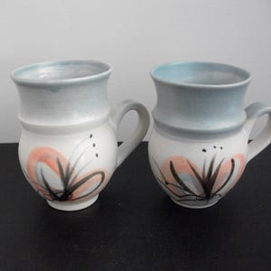 May include: Two ceramic mugs with a white base, light blue rim, and a curved handle. Each mug features a floral design in orange, black, and grey. The mugs are displayed on a dark surface against a white background.
