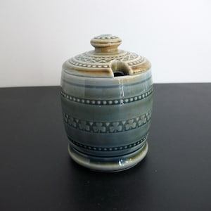 May include: A ceramic sugar bowl with a lid, featuring a blue-green glaze and decorative patterns. The bowl has a cylindrical shape with horizontal bands of design, including a clover pattern. The lid has a knob and a spoon slot.