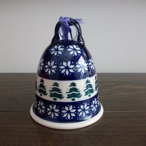 May include: A ceramic bell with a dark blue glaze, decorated with white snowflake and green Christmas tree patterns. A purple ribbon is attached to the top. The bell is on a wooden surface.
