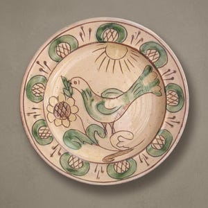 May include: A round ceramic plate with a cream-coloured base featuring a hand-painted green bird holding a leaf, a yellow flower, and a sun motif in the centre. The rim is decorated with a repeating pattern of green circles with cross-hatching.