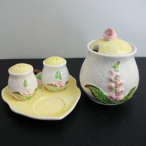 May include: A ceramic salt and pepper set and sugar bowl. The set includes a yellow leaf-shaped tray, two salt and pepper shakers, and a sugar bowl with a lid. All pieces are white with floral and leaf designs, suitable for the kitchen.