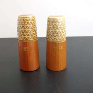 May include: A pair of vintage salt and pepper shakers. They feature a wooden base and a cream-coloured top with a geometric pattern. The wooden base is light brown and has a small yellow logo.