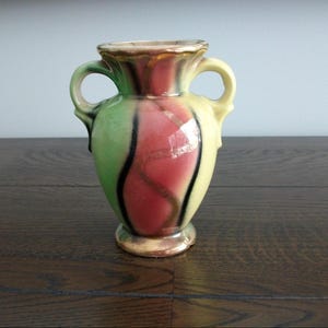 May include: A ceramic vase featuring two handles and a flared rim. The body displays a vertical colour pattern of light green, soft pink, and yellow, accented by dark black wavy lines and a metallic gold trim along the rim and base edges.