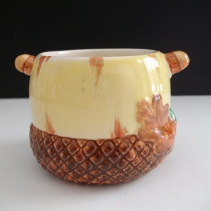 May include: A ceramic acorn-shaped container with a yellow glaze and brown accents. The container has a textured brown base and a leaf design on the side.