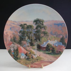 May include: A vintage ceramic plate featuring a painted image of a rural landscape. The scene depicts a small village with houses, trees, and a dirt road. The plate has a light blue border and the text "C.E.Wallis Artist" is printed in the bottom right corner.