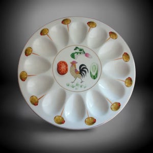 May include: A white ceramic serving platter with 12 oval indentations for appetizers. The centre features a rooster illustration with vegetables. Olives on sticks are arranged around the edge. A decorative and functional piece for entertaining.