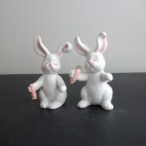 May include: Two white ceramic rabbit figurines with pink inner ears and closed eyes, each holding a carrot. The rabbits have a glossy finish and are posed as if they are dancing or playing.