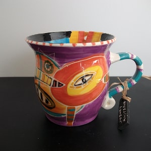May include: A hand-painted ceramic mug with a vibrant, abstract design. The mug features a purple body with orange, yellow, and blue accents. The handle is striped with turquoise and white. A tag is tied to the handle.