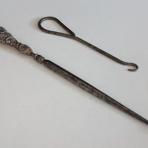 May include: Two antique metal tools on a white surface. One tool has a decorative handle and a hook at the end. The other tool is a simple metal hook. Both tools appear to be vintage and show signs of wear.