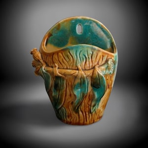 May include: A ceramic wall pocket planter featuring a textured brown tree bark finish with applied teal green leaves and a branch detail. The vessel has a rounded opening and a single hole at the top for mounting, with a glossy glazed surface.