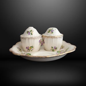 May include: A ceramic salt and pepper shaker set on a matching oval tray. The off-white pieces feature a delicate purple violet floral pattern, vertical ribbing, and scalloped edges with a worn gold-tone trim along the rims and base.