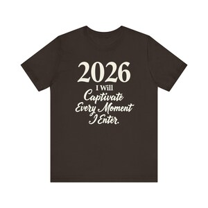 Puede incluir: Una camiseta marrón oscuro con el año "2026" impreso en blanco, en negrita y en gran tamaño. Debajo, el texto "I Will Captivate Every Moment I Enter" está escrito en una fuente cursiva elegante. La camiseta tiene mangas cortas y cuello redondo.