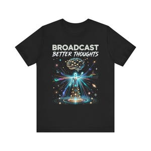May include: A black t-shirt featuring the white text BROADCAST BETTER THOUGHTS above a graphic of a glowing human silhouette standing on a transmission tower, surrounded by colourful light rays, stars, and a floating brain with connected nodes.
