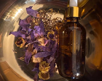 May include: A small amber glass dropper bottle with a gold collar and white bulb, positioned inside a polished gold-toned metal bowl alongside a collection of dried purple flower petals and small yellow botanical elements.