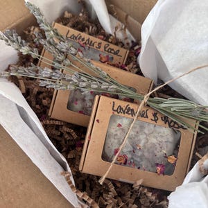 Puede incluir: Una caja de regalo de cartón marrón que contiene dos pequeñas cajas de jabón, etiquetadas como "Lavender & Rose". Las cajas están llenas de jabón y pétalos de rosa secos. La caja está forrada con papel blanco y papel triturado marrón, y decorada con ramitas de lavanda.