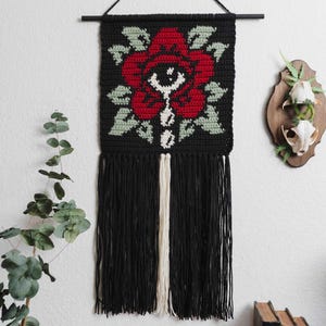 May include: Black crochet wall hanging featuring a red rose design with green leaves and a white accent. The wall hanging has black fringe and a wooden rod for hanging. Decorative wall art.