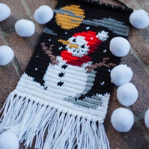 Könnte beinhalten: Handgefertigter Wandbehang aus Häkelarbeit mit einem Schneemann-Design. Der Schneemann trägt einen roten Hut und Schal vor einem schwarzen Hintergrund mit gelbem Mond und fallendem Schnee. Weiße Fransen hängen von unten. Weiße Pompons sind verstreut.