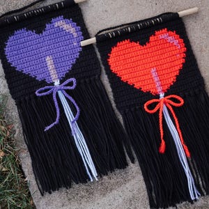 May include: Two crochet wall hangings with black backgrounds and heart-shaped lollipops. One features a purple heart, the other a red heart. Each has a wooden dowel, fringe, and a bow. The lollipops have white and purple or red and white sticks.