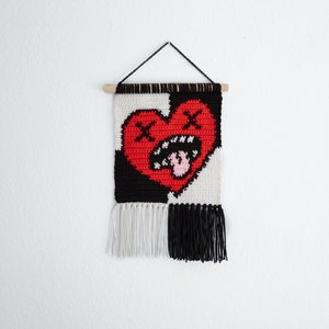 Puede incluir: Un tapiz tejido en blanco y negro con un coraz&oacute;n rojo con una cara fruncida y una lengua rosa que sobresale. El coraz&oacute;n tiene dos X sobre &eacute;l. El tapiz tiene flecos blancos y negros en la parte inferior.