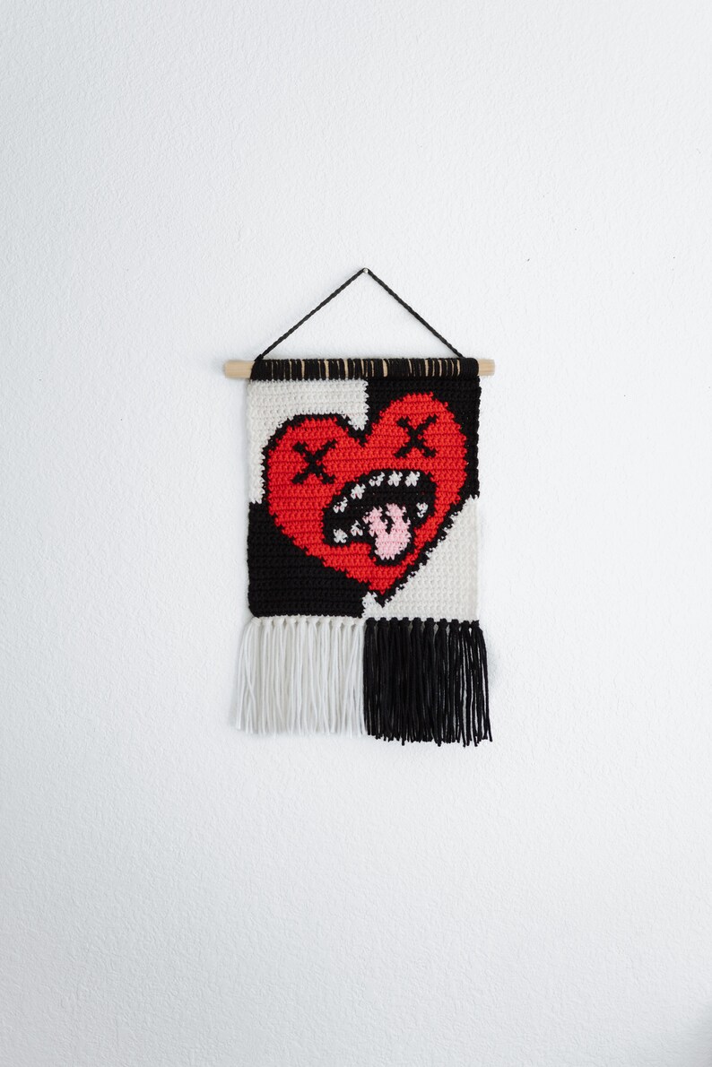 Puede incluir: Un tapiz tejido en blanco y negro con un coraz&oacute;n rojo con una cara fruncida y una lengua rosa que sobresale. El coraz&oacute;n tiene dos X sobre &eacute;l. El tapiz tiene flecos blancos y negros en la parte inferior.