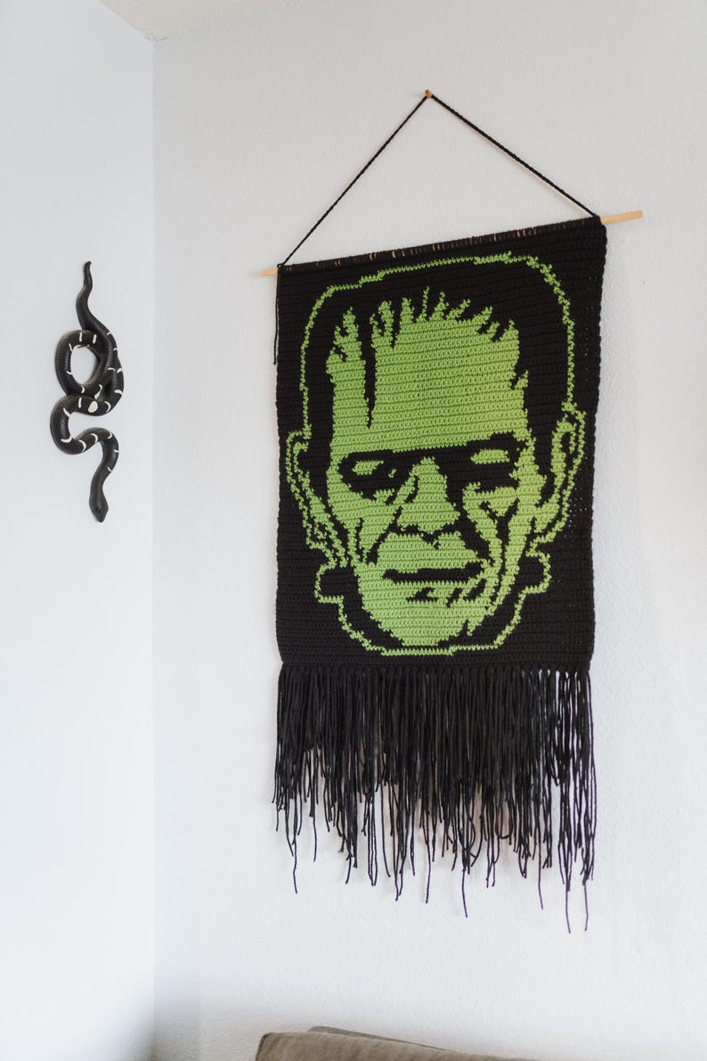 May include: A black and green crocheted wall hanging featuring a green Frankenstein face with black eyes and a black fringe border.