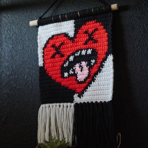 Puede incluir: Un tapiz de pared de crochet blanco y negro con un coraz&oacute;n rojo en el centro. El coraz&oacute;n tiene un contorno negro y una boca blanca con dientes rosados. El tapiz tiene flecos blancos y negros en la parte inferior.