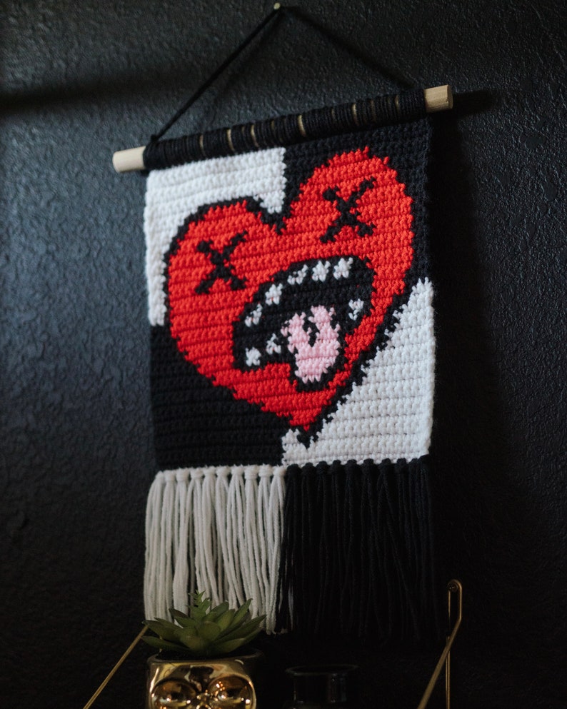 Puede incluir: Un tapiz de pared de crochet blanco y negro con un coraz&oacute;n rojo en el centro. El coraz&oacute;n tiene un contorno negro y una boca blanca con dientes rosados. El tapiz tiene flecos blancos y negros en la parte inferior.