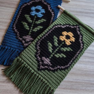 May include: Two handmade crochet wall hangings. One is blue, the other green. Each features a black floral design with a flower and leaves. The hangings have fringe at the bottom and are mounted on wooden dowels.