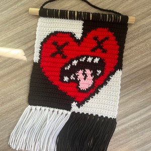Puede incluir: Un tapiz de pared tejido a crochet con un coraz&oacute;n rojo con contorno negro y una lengua rosa que sobresale. El coraz&oacute;n tiene dos X negras para los ojos. El fondo es negro y blanco con flecos blancos a la izquierda y flecos negros a la derecha.