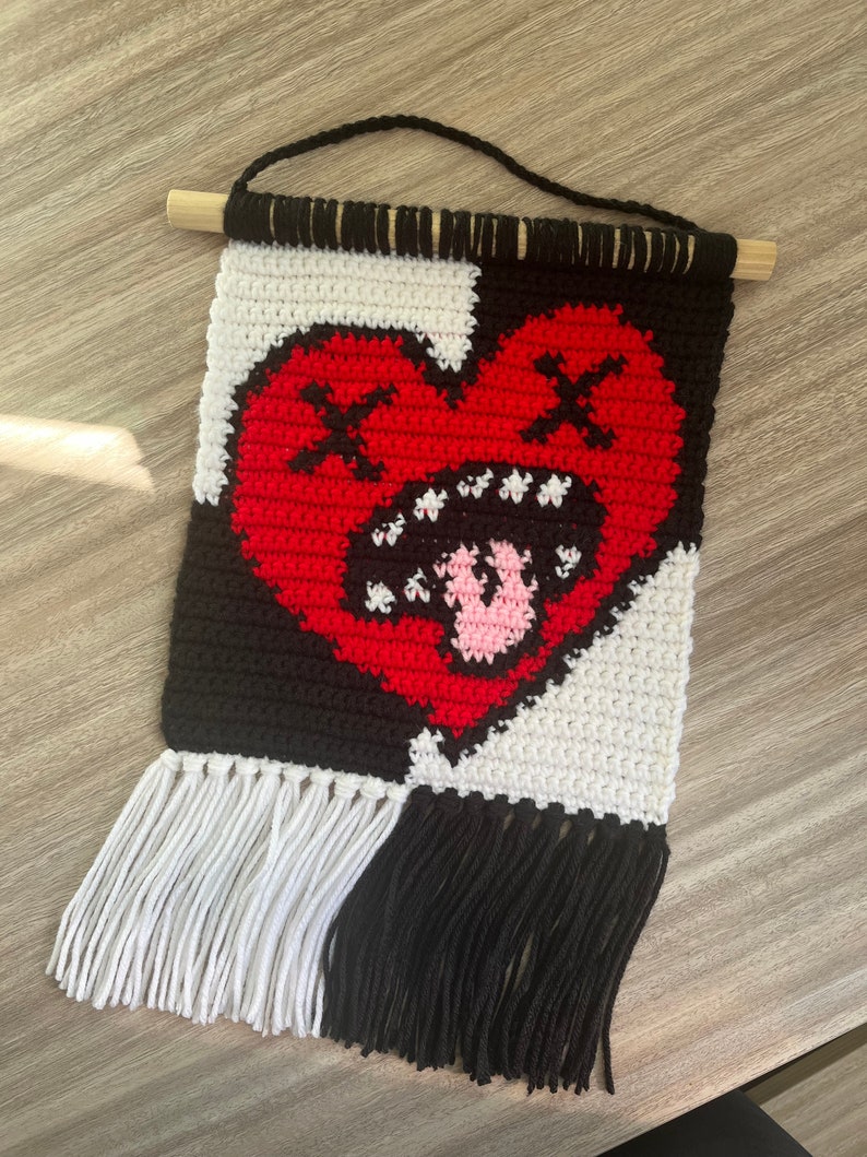 Puede incluir: Un tapiz de pared tejido a crochet con un coraz&oacute;n rojo con contorno negro y una lengua rosa que sobresale. El coraz&oacute;n tiene dos X negras para los ojos. El fondo es negro y blanco con flecos blancos a la izquierda y flecos negros a la derecha.