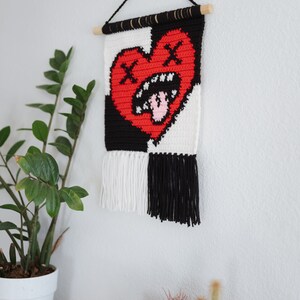 Puede incluir: Un tapiz de pared de crochet blanco y negro con un coraz&oacute;n rojo en el centro. El coraz&oacute;n tiene un aspecto desgastado con una cara fruncida y una lengua rosa que sobresale. El tapiz tiene flecos blancos y negros en la parte inferior.