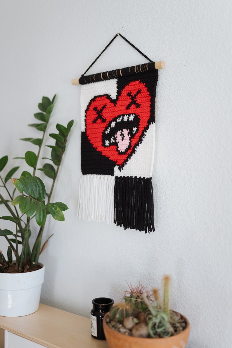 Puede incluir: Un tapiz de pared de crochet blanco y negro con un coraz&oacute;n rojo en el centro. El coraz&oacute;n tiene un aspecto desgastado con una cara fruncida y una lengua rosa que sobresale. El tapiz tiene flecos blancos y negros en la parte inferior.