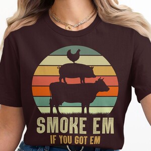 Puede incluir: Una camiseta marrón con un gráfico de estilo retro de un pollo, un cerdo y una vaca apilados uno encima del otro. El gráfico está sobre un fondo de puesta de sol colorido. El texto "Smoke Em If You Got Em" está impreso debajo del gráfico.