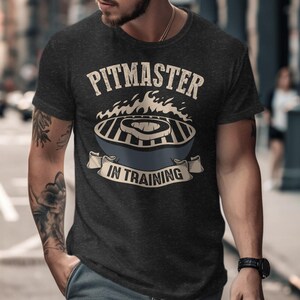 May include: A dark grey t-shirt with a graphic of a grill and flames. The text "Pitmaster in Training" is printed on the shirt.