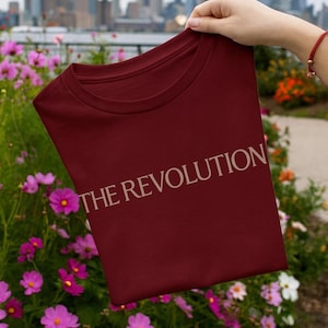 Puede incluir: Una camiseta granate con el texto "THE REVOLUTION" en color beige claro. La camiseta se sostiene frente a un paisaje urbano con un cielo nublado y un jardín de flores en primer plano.