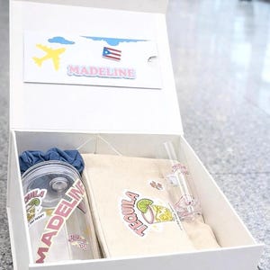 Puede incluir: Una caja de regalo blanca que contiene un juego de regalo personalizado con temática de viaje. El juego incluye un vaso transparente con el nombre "Madeline", una bolsa de lona con un gráfico de tequila y una pajita de vidrio pequeña. También se incluye una tarjeta con un avión, nubes y la bandera puertorriqueña.