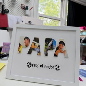 Puede incluir: Un marco de fotos rectangular blanco que contiene la palabra PAPA, donde cada letra está rellena con una fotografía diferente de un padre y un hijo. Debajo de la palabra, el texto Eres el mejor está impreso entre dos iconos de balón.