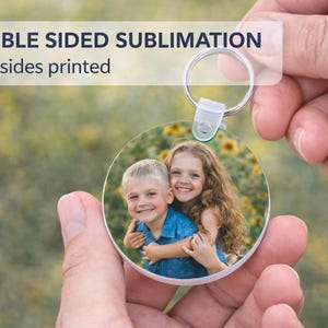 Könnte beinhalten: Ein runder Schlüsselanhänger mit einem Foto von zwei lächelnden Kindern. Der Schlüsselanhänger hat einen silbernen Ring und einen weißen Kunststoffverschluss. Der Text "DOUBLE SIDED SUBLIMATION Both sides printed" steht oben.