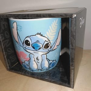 May include: A light blue ceramic mug featuring the character Stitch from Disney's Lilo & Stitch. The mug is decorated with a smiling Stitch, pink flowers, and green leaves. The mug is packaged in a black box with the Disney logo.