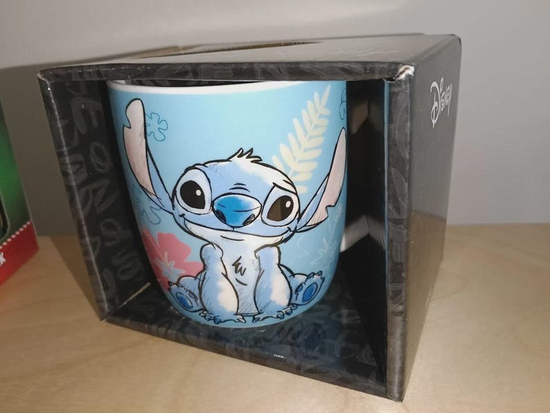 May include: A light blue ceramic mug featuring the character Stitch from Disney's Lilo & Stitch. The mug is decorated with a smiling Stitch, pink flowers, and green leaves. The mug is packaged in a black box with the Disney logo.