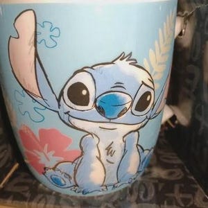 May include: A light blue ceramic mug featuring a cartoon illustration of Stitch from Lilo & Stitch. The mug has a wraparound design with Stitch's face, a pink flower, and a leafy pattern. The mug is in a box.