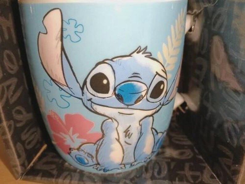 May include: A light blue ceramic mug featuring a cartoon illustration of Stitch from Lilo & Stitch. The mug has a wraparound design with Stitch's face, a pink flower, and a leafy pattern. The mug is in a box.