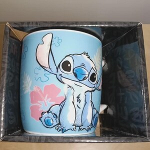 May include: A light blue ceramic mug featuring the character Stitch from Lilo & Stitch. The mug has a cartoon illustration of Stitch, a pink hibiscus flower, and other floral designs. The mug is packaged in a black box.