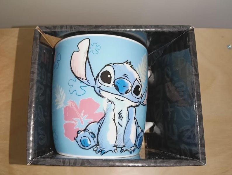 May include: A light blue ceramic mug featuring the character Stitch from Lilo & Stitch. The mug has a cartoon illustration of Stitch, a pink hibiscus flower, and other floral designs. The mug is packaged in a black box.