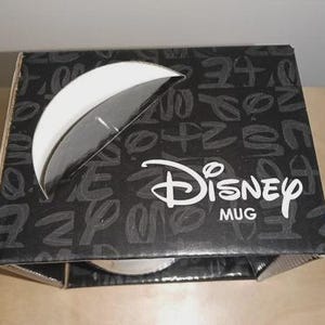 May include: A black and white Disney mug box with a partial view of the mug inside. The box has a white semi-circle cutout and the Disney logo in white. The box is covered in a pattern of white letters.